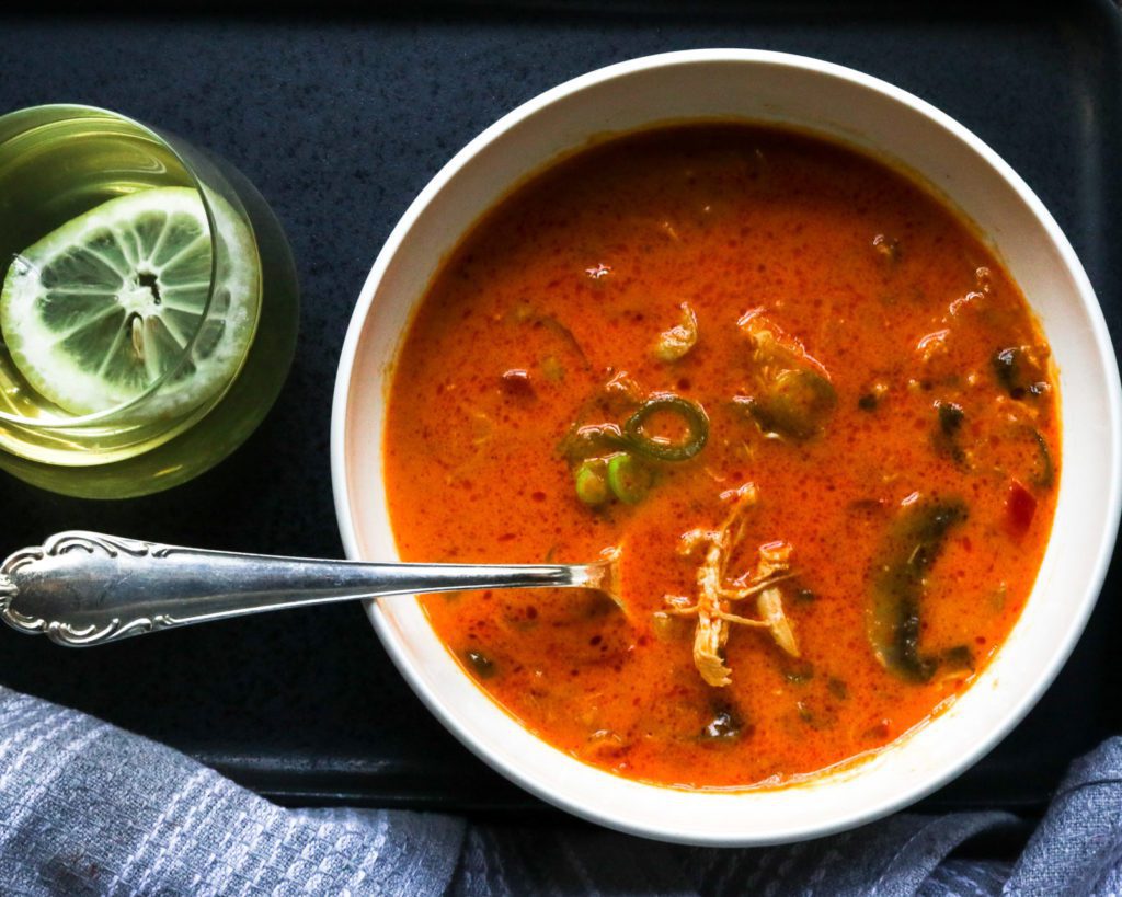 Tom Kha Gai Chicken Soup in white bowl, glass with liquid and lemon, dark background