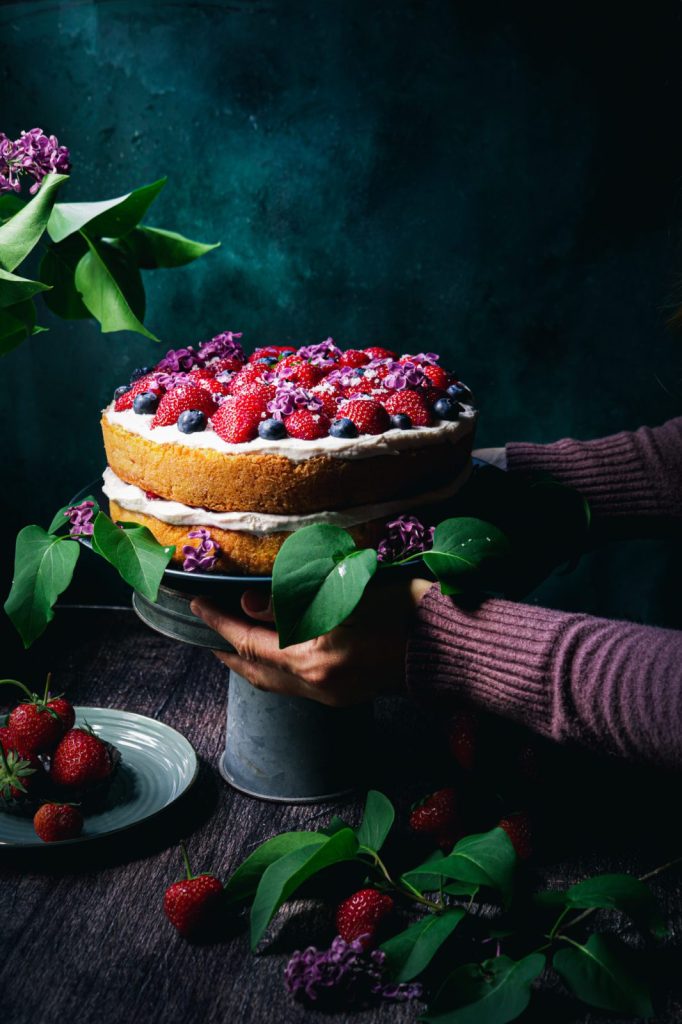  cake with lemon cream cheese frosting and berries on a dark plate with hands in frame