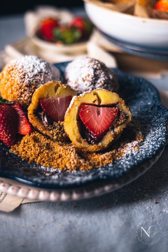 topfenknödel- Austrian cheese curd dumplings cut in half filled with a strawberry and Nutella, on a blue plate with bread crumbs and powdered sugar